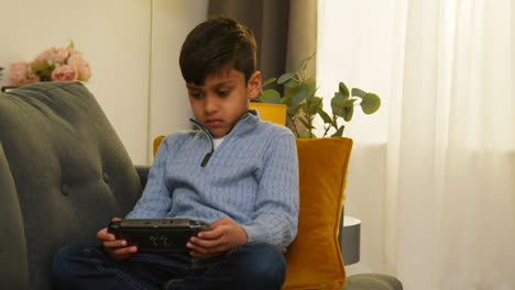 Young-Boy-Sitting-On-Sofa-At-Home-Playing-Game-Or-Streaming-Onto-Handheld-Gaming-Device